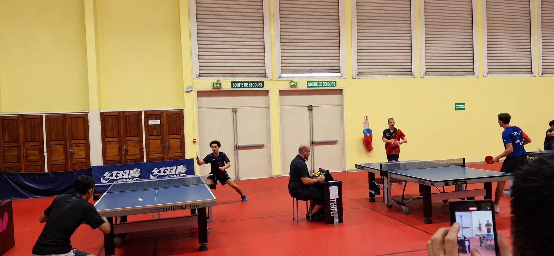 Vue d’ensemble de deux rencontres de tennis de table avec un spectateur filmant au premier plan